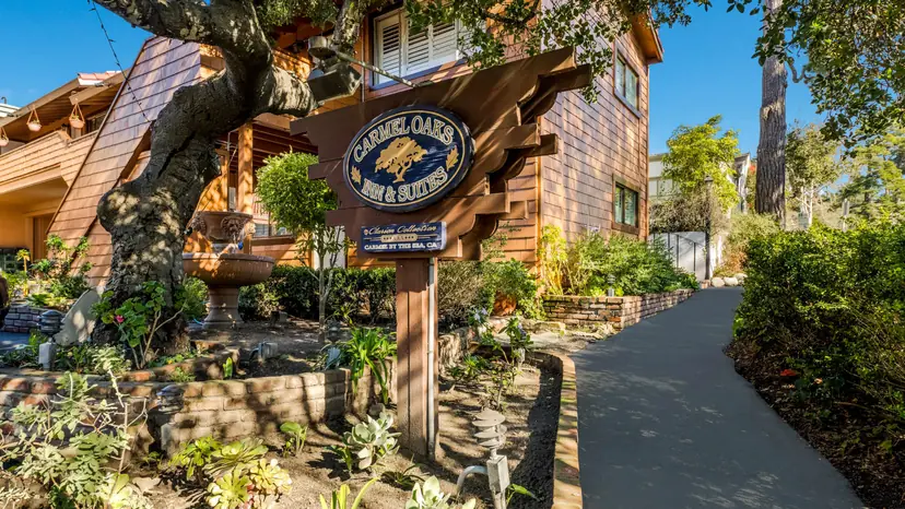 The carved Carmel Oaks Inn & Suites sign, navy with a gold oak tree, beside an old oak, a stone fountain and the inn's cedar-shingled walls