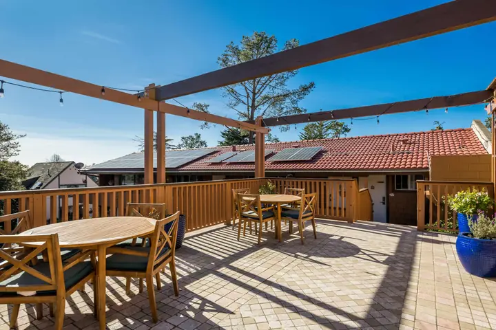 Sun deck under a wooden pergola with string lights, teak tables and chairs and a blue planter